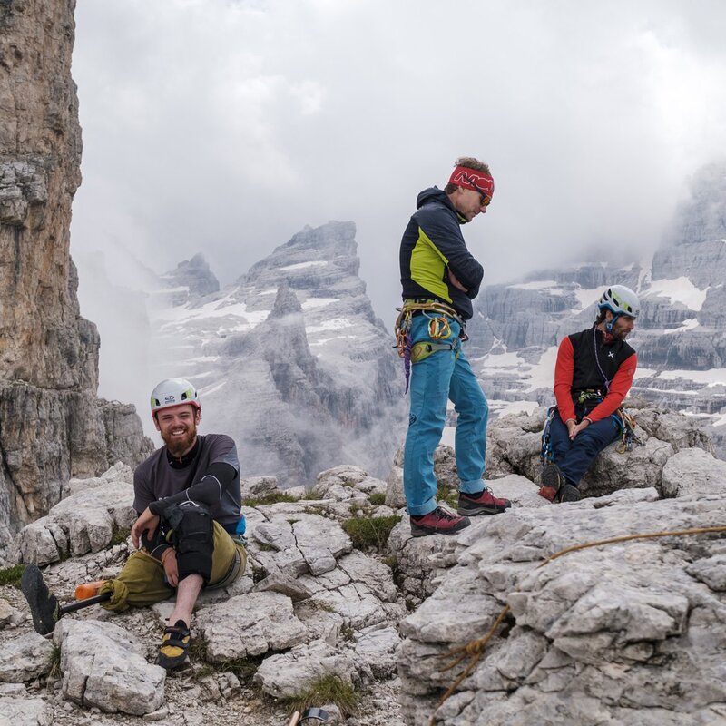 Tre uomini equipaggiati con caschi e imbracature da arrampicata si trovano su una cengia rocciosa in un ambiente di alta montagna, caratterizzato da imponenti pareti e cime rocciose parzialmente avvolte dalle nuvole. L'uomo seduto a sinistra, che sorride alla camera, indossa una protesi alla gamba sinistra. | © Filippo Frizzera, 2021