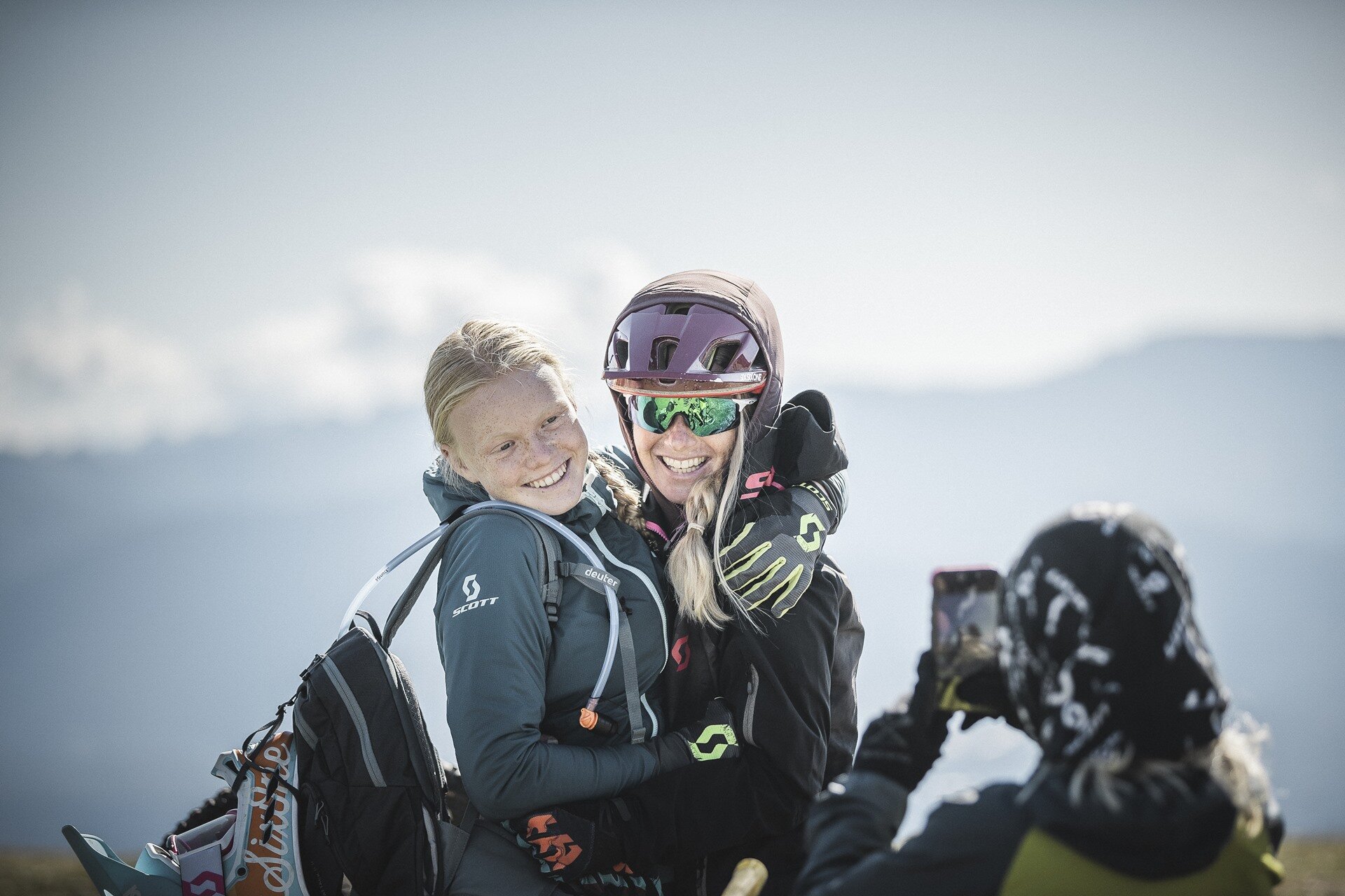 Due giovani ragazze adolescenti si abbracciano e sorridono mentre una terza persona scatta loro una foto. Sono vestite da biker, con guanti, protezioni e casco. | © Manuel Kottersteger, 2020