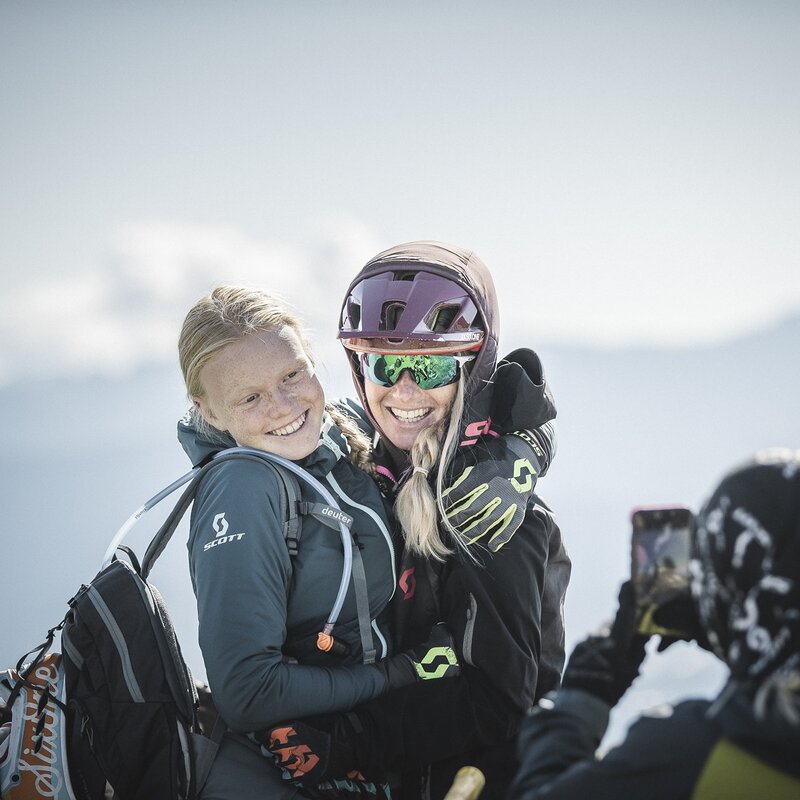 Due giovani ragazze adolescenti si abbracciano e sorridono mentre una terza persona scatta loro una foto. Sono vestite da biker, con guanti, protezioni e casco. | © Manuel Kottersteger, 2020