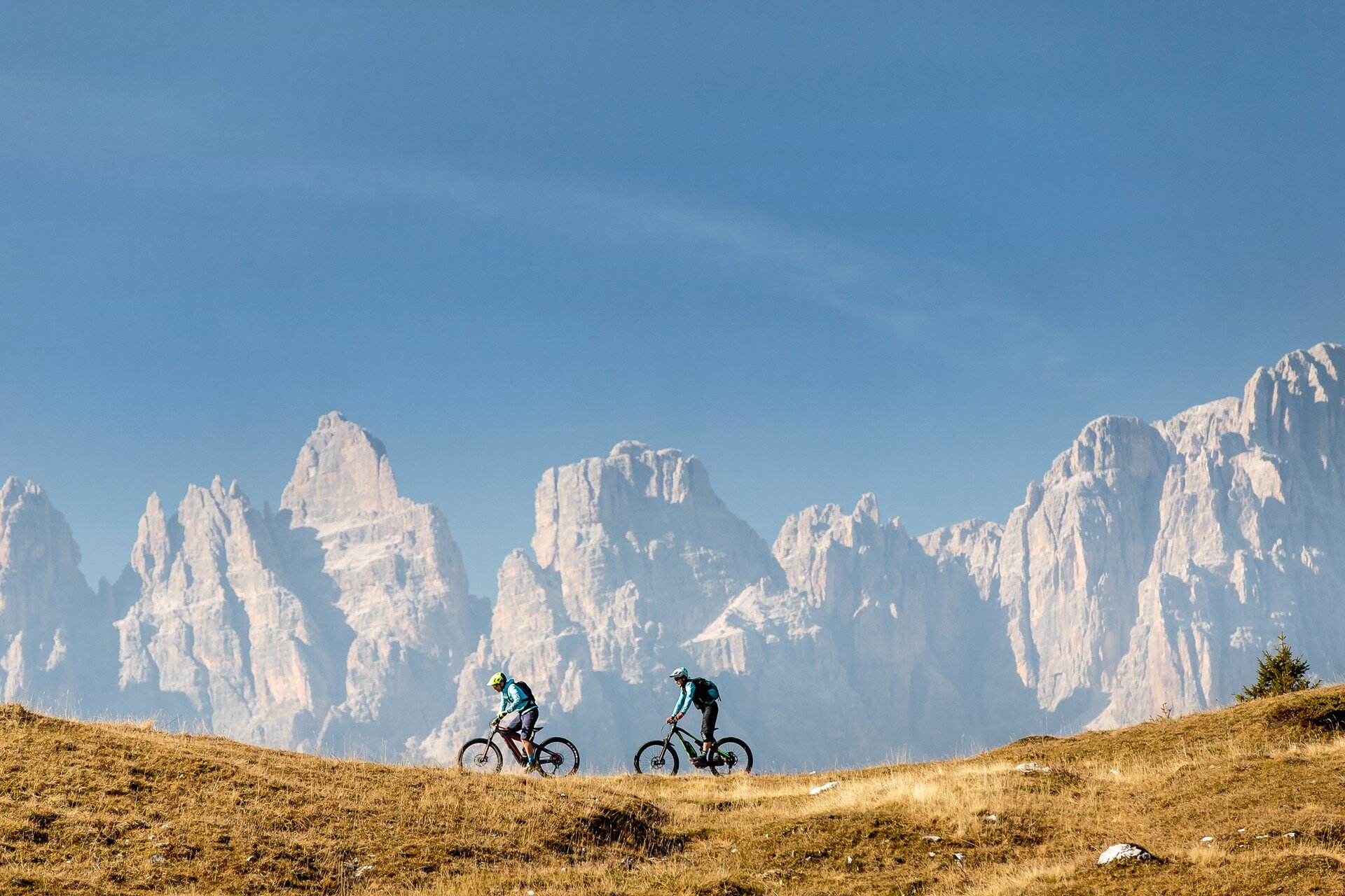 Due ciclisti con casco e abbigliamento tecnico percorrono un sentiero erboso in e-MTB. Sullo sfondo, le imponenti cime rocciose delle Dolomiti di Brenta si stagliano contro un cielo limpido e azzurro. L'erba in primo piano ha tonalità dorate, suggerendo una stagione autunnale. | © Alex Luise, 2017