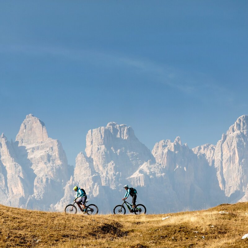 Due ciclisti con casco e abbigliamento tecnico percorrono un sentiero erboso in e-MTB. Sullo sfondo, le imponenti cime rocciose delle Dolomiti di Brenta si stagliano contro un cielo limpido e azzurro. L'erba in primo piano ha tonalità dorate, suggerendo una stagione autunnale. | © Alex Luise, 2017