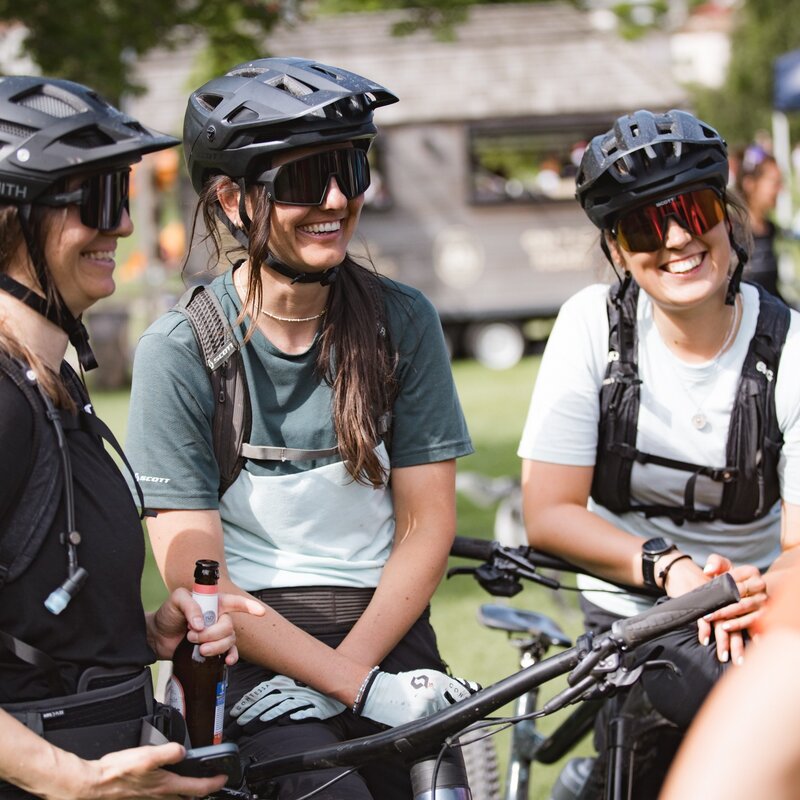 Tre donne sorridenti, equipaggiate con caschi da bicicletta, occhiali da sole e zainetti, sono in pausa durante un evento ciclistico all'aperto. Una di loro tiene una bevanda, mentre le altre due sono vicine alle loro biciclette, in un'atmosfera allegra e conviviale. | © Mia Knoll, 2024