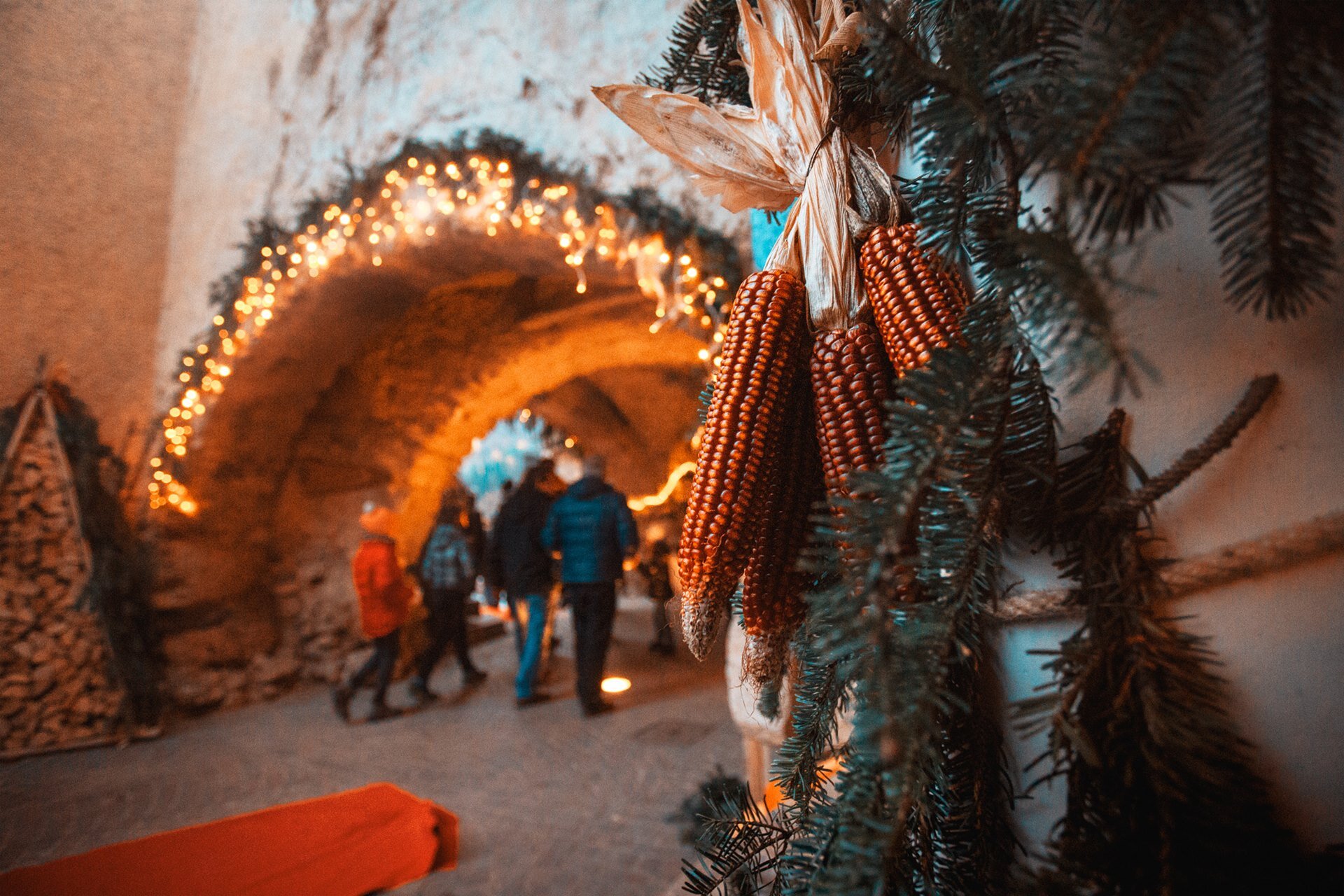 In the foreground, some deep red cobs of corn hang from a fir tree branch. In the background, one can see a stone arch illuminated by small warm lights, forming a bright garland. Some people, dressed in winter clothes, walk under the arch. The contrast between the lights and the old architecture gives the image a cosy and traditional Christmas atmosphere. | © Filippo Frizzera, 2018