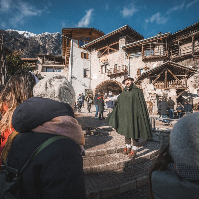 This photo shows a man dressed in a dark green cloak and hat who is leading or entertaining a group of people outside the village of Rango. In the background can be seen stone houses with wooden balconies and, in the distance, a snow-capped mountain. Some tourists wear heavy jackets and caps to protect themselves from the cold as they watch the man, who smiles and seems to be talking. | © Archivio Garda Dolomiti Spa