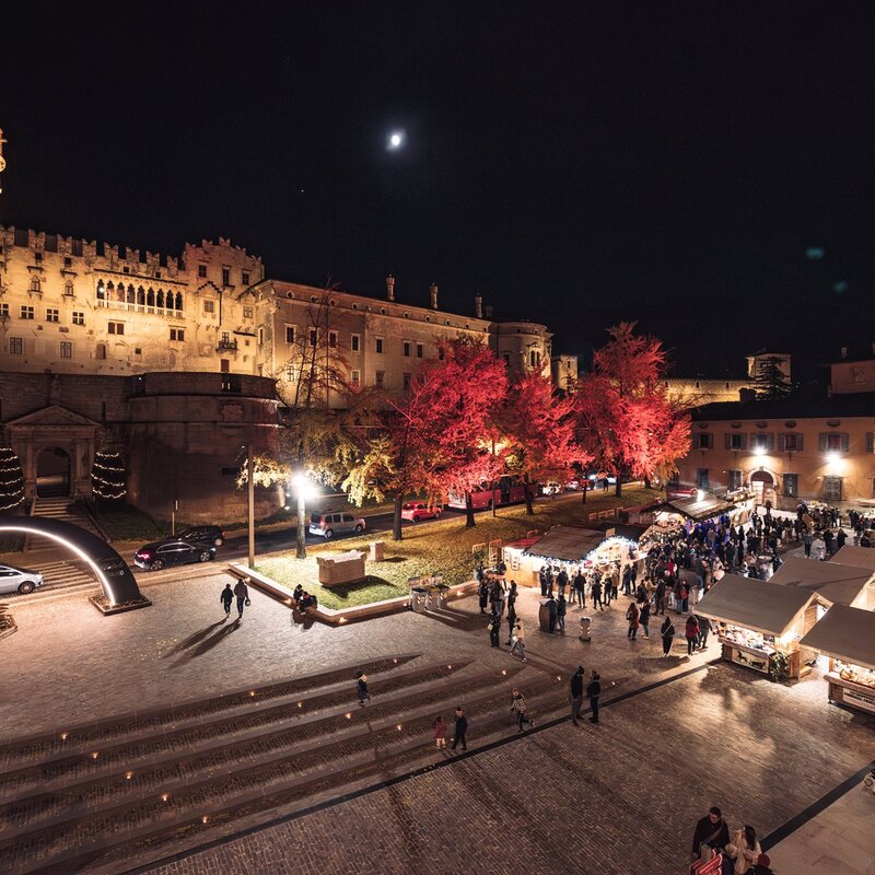 This picture shows the night-time Trento Christmas Market in the square in front of the Buonconsiglio Castle. The castle, illuminated by warm lights, has an imposing structure with a circular tower on the left and crenellated walls. The trees along the square are decorated with red lights that make them stand out in the darkness. The market stalls are arranged in neat rows, each with a small white roof, and are internally lit, creating a cosy and festive atmosphere. Several people stroll among the stalls, enjoying the event. In the night sky, a small moon is visible above the castle. | © M. Gober - Archivio APT Trento
