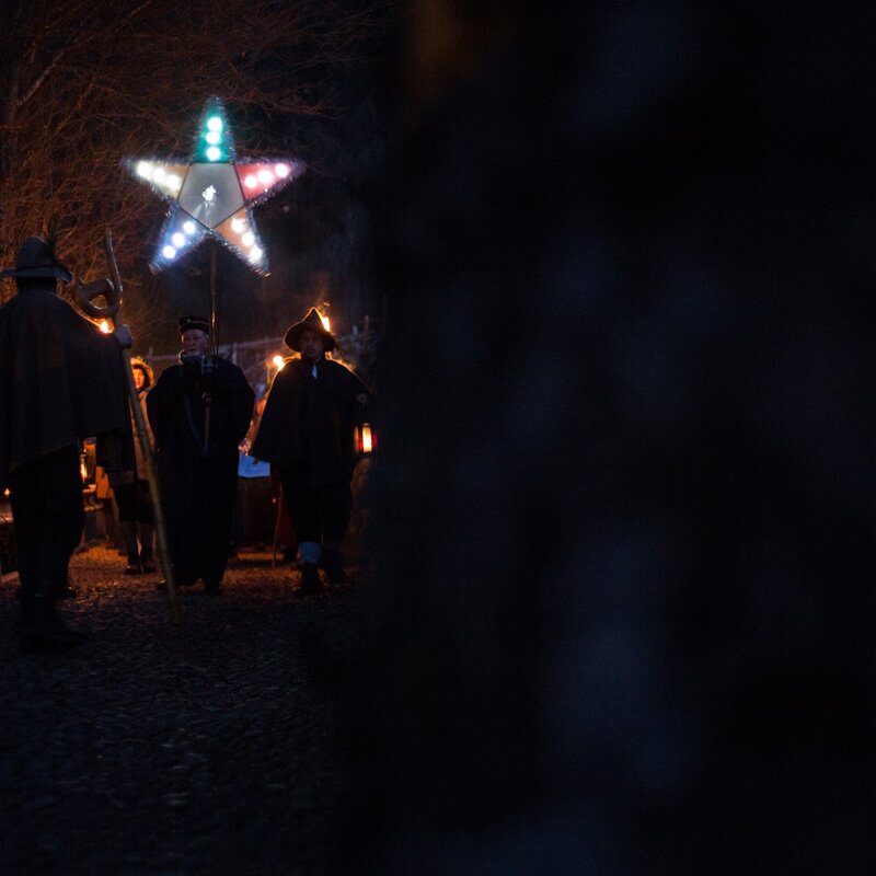 A group of people walk along a cobbled path at night, carrying lighted torches and a large, multicoloured star on a pole. In the background, bare trees can be glimpsed, faintly illuminated by torchlight. | © Alfredo Croce, 2017