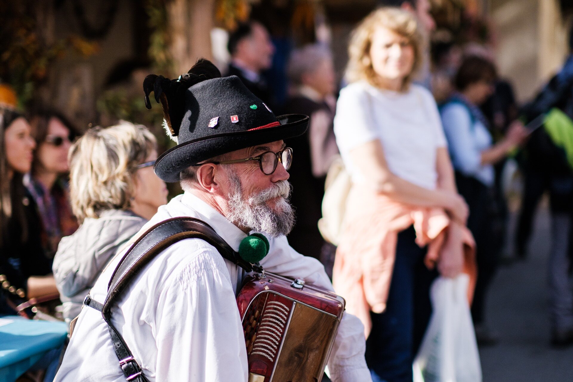 A musician with a beard and glasses, wearing a traditional hat, plays the accordion during the Sagra della Ciuìga festival in San Lorenzo Dorsino. The close-up captures the artist in folkloric attire, immersed in the atmosphere of a lively village festival, with the audience visible and out of focus in the background. | © Valerio Rigotti, 2022