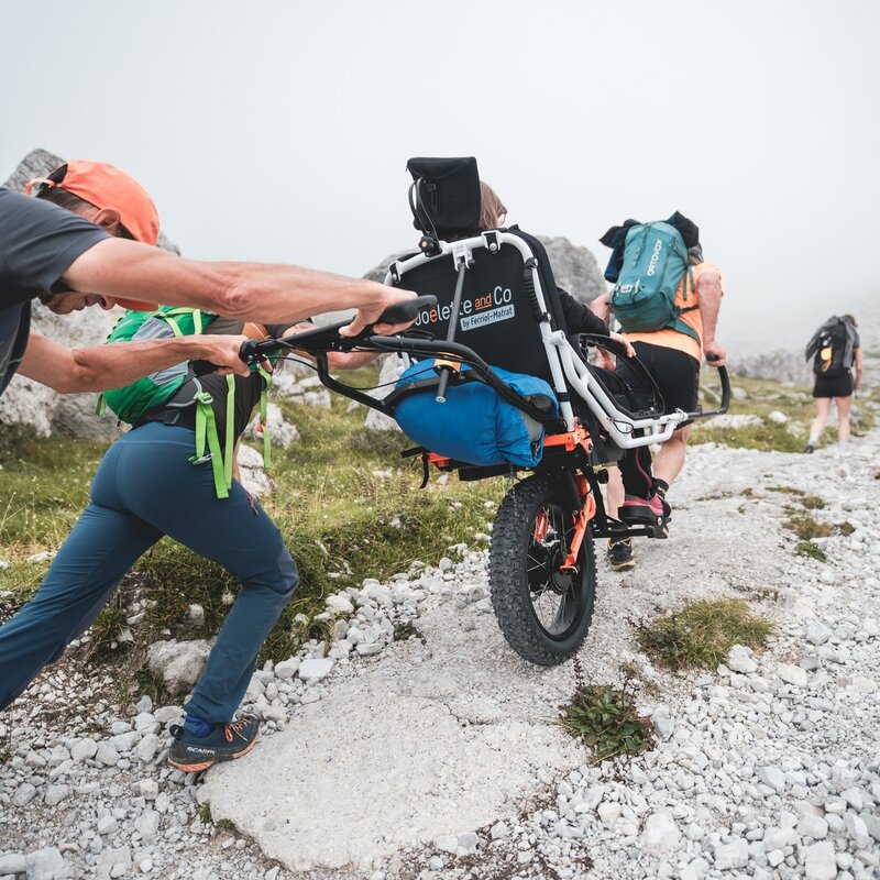 Due escursionisti trasportano una persona seduta su una Joëlette, una speciale carrozzina monoruota progettata per il trekking inclusivo, lungo un ripido sentiero roccioso di montagna. La scena evidenzia il lavoro di squadra: un accompagnatore tira la struttura dalla parte anteriore mentre l'altro spinge da dietro per superare la pendenza. Sullo sfondo, avvolto da una leggera nebbia, altri escursionisti con zaini proseguono il cammino in un paesaggio d'alta quota, sulle Dolomiti di Brenta. | © Filippo Frizzera, 2025