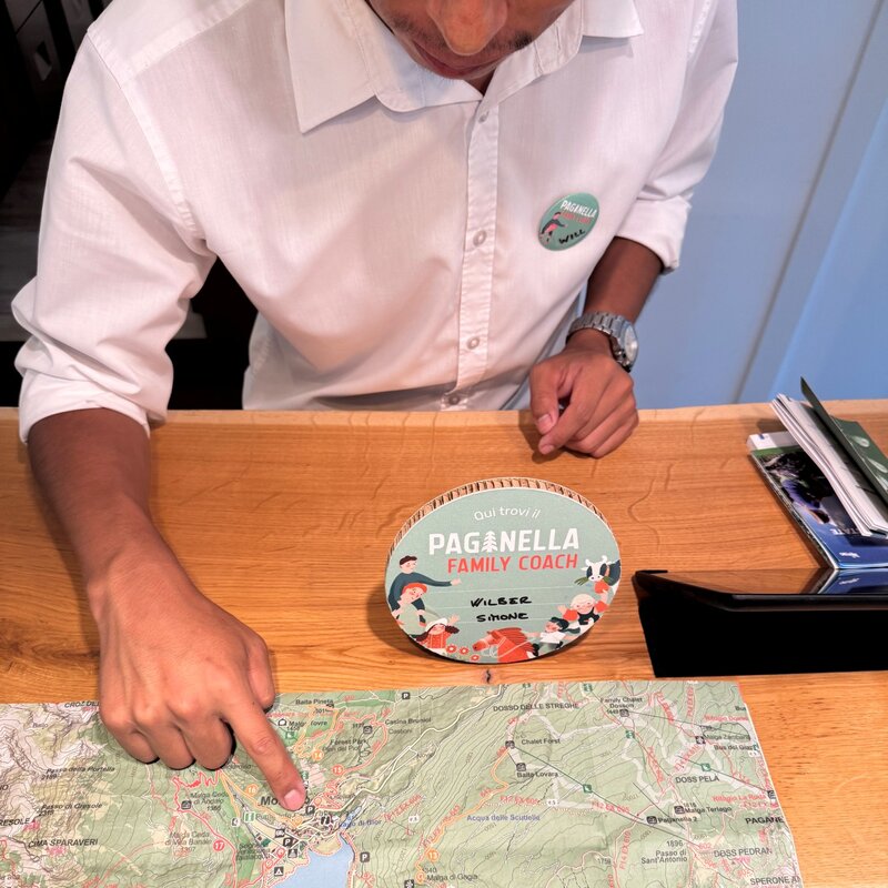 Una persona, che indossa una camicia bianca e una spilla identificativa, indica con l'indice un punto preciso su una mappa escursionistica stesa su un tavolo di legno. Davanti a lei è posizionato un cartello rotondo in cartone che recita: "Qui trovi il PAGANELLA FAMILY COACH", con scritti a mano i nomi "WILBER" e "SIMONE". | © APT Paganella, 2025