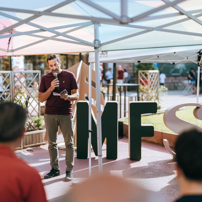 Un relatore parla al microfono consultando degli appunti, in piedi sotto un gazebo bianco durante un evento all'aperto ad Andalo. Alle sue spalle si notano le grandi lettere verdi "MFS", simbolo del festival Mountain Future Stories, mentre il pubblico ascolta in primo piano. | © Mirko Perli, 2025