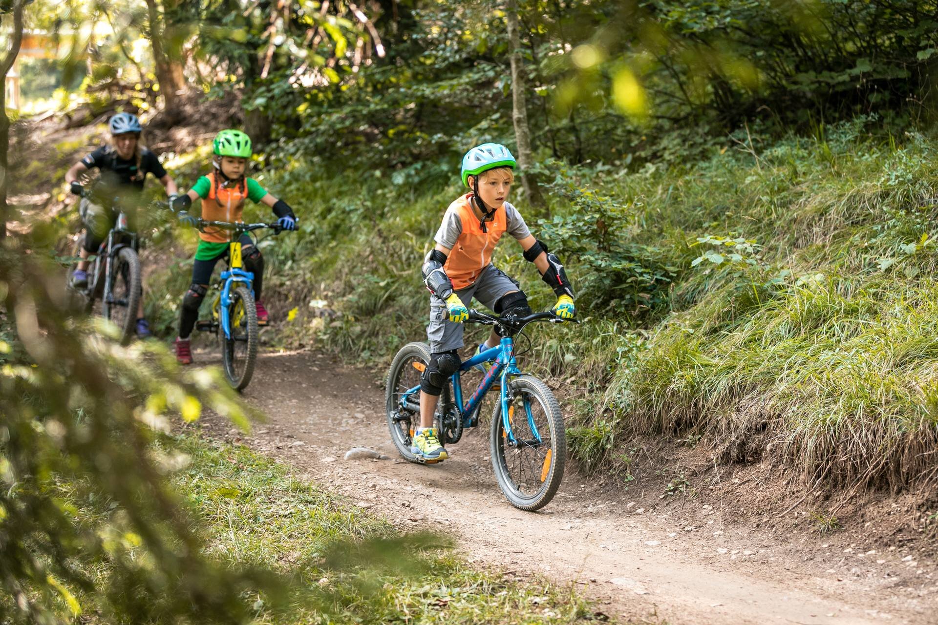 Zwei Kinder und ein Fahrradführer der Akademie Paganella radeln einen Waldweg hinunter. Sie tragen Helme und Protektoren.  | © Oliver Astrologo, 2019