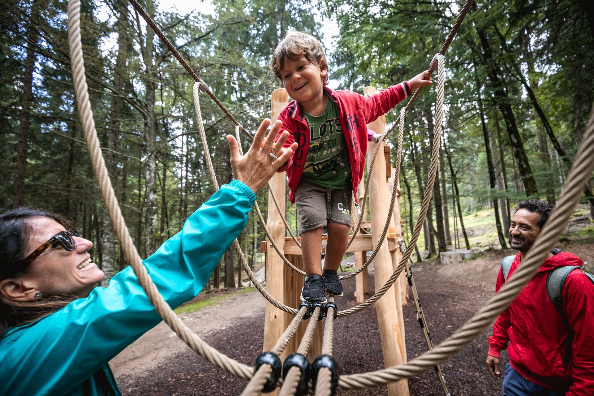 A small child walks on a Tibetan bridge made of ropes and wooden planks, inside a forest. He wears a green T-shirt, a red sweatshirt and short shorts. He holds out his hand to give a ‘high five’ to a woman who looks up at him from the ground with a smile, wearing a turquoise jacket and sunglasses. A man in a red jacket and backpack watches the scene with a smile, on the opposite side. Tall, dense trees surround the area.  Translated with DeepL.com (free version) | © Oliver Astrologo, 2019