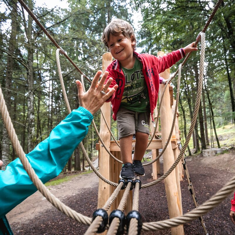 Un bambino piccolo cammina su un ponte tibetano fatto di corde e assi di legno, all’interno di un bosco. Indossa una maglietta verde, una felpa rossa e pantaloncini corti. Tende la mano per dare un “cinque” a una donna che lo guarda da terra con un sorriso, indossando una giacca turchese e occhiali da sole. Un uomo con giacca rossa e zaino osserva la scena sorridendo, sul lato opposto. Gli alberi alti e fitti circondano l’area. | © Oliver Astrologo, 2019