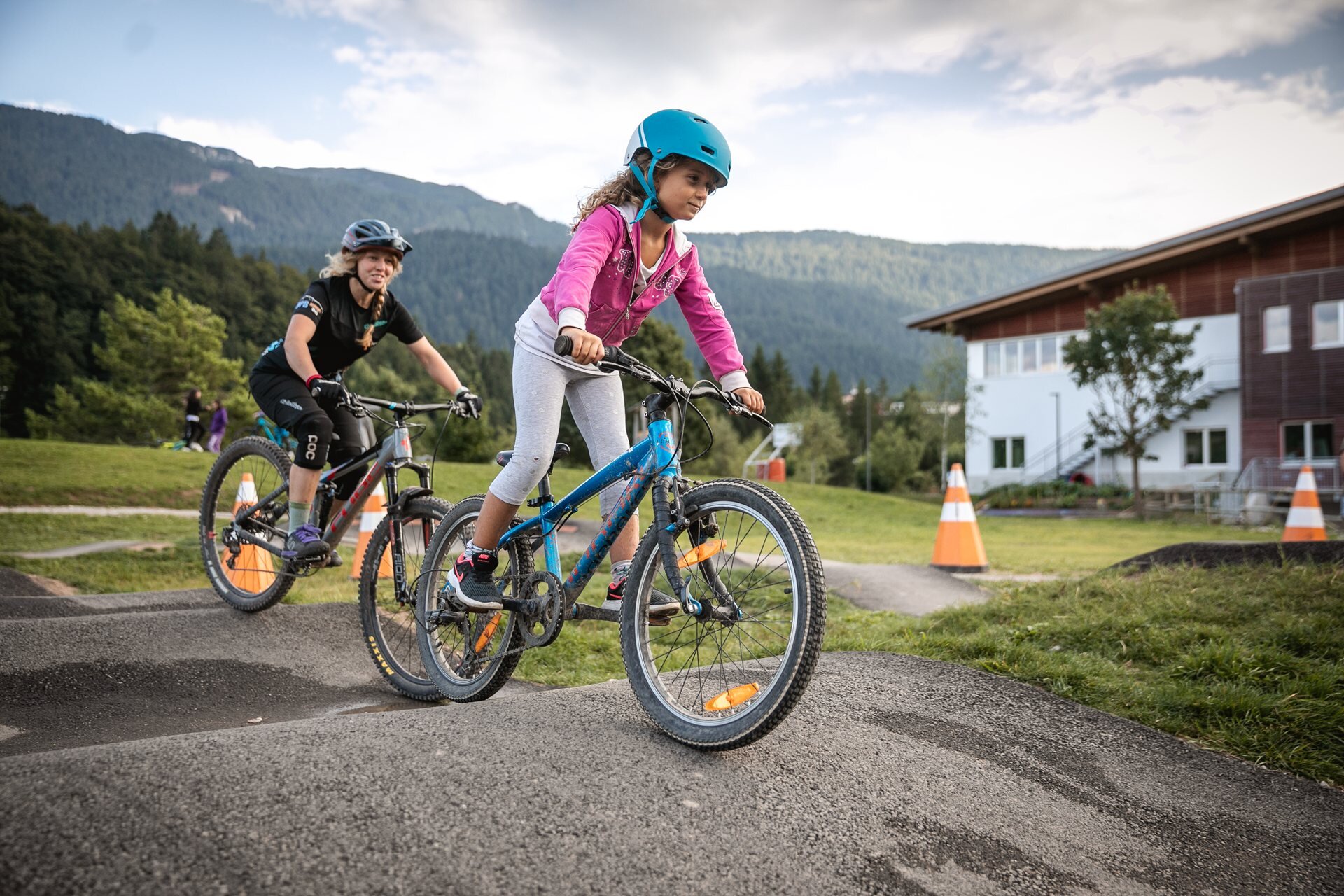 Due persone in bicicletta stanno percorrendo un pump track in un ambiente montano. La bambina in primo piano indossa un casco azzurro, una maglia rosa e pantaloni grigi, mentre la donna dietro, insegnante bike, ha un casco grigio, una maglia nera e pantaloncini neri. Il percorso è delimitato da coni arancioni e sullo sfondo si vedono montagne e un edificio chiaro. | © Oliver Astrologo, 2019