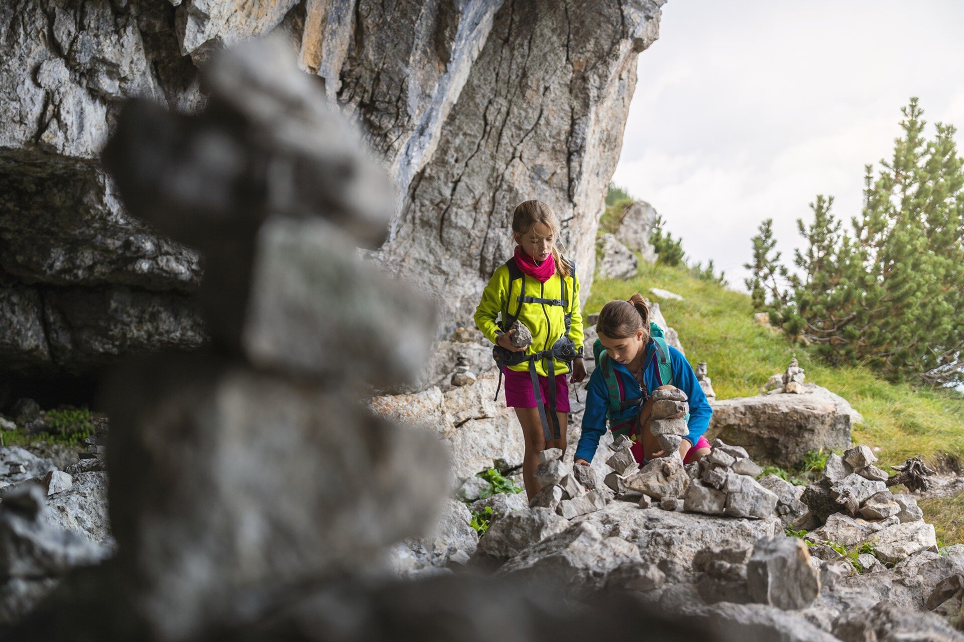 Due bambine sono intente a creare delle torri con piccole pietre. Una è accovacciata e sistema i sassi, mentre l'altra la osserva in piedi, vicino all'apertura di una grotta e con abeti sullo sfondo. | © Filippo Frizzera, 2019