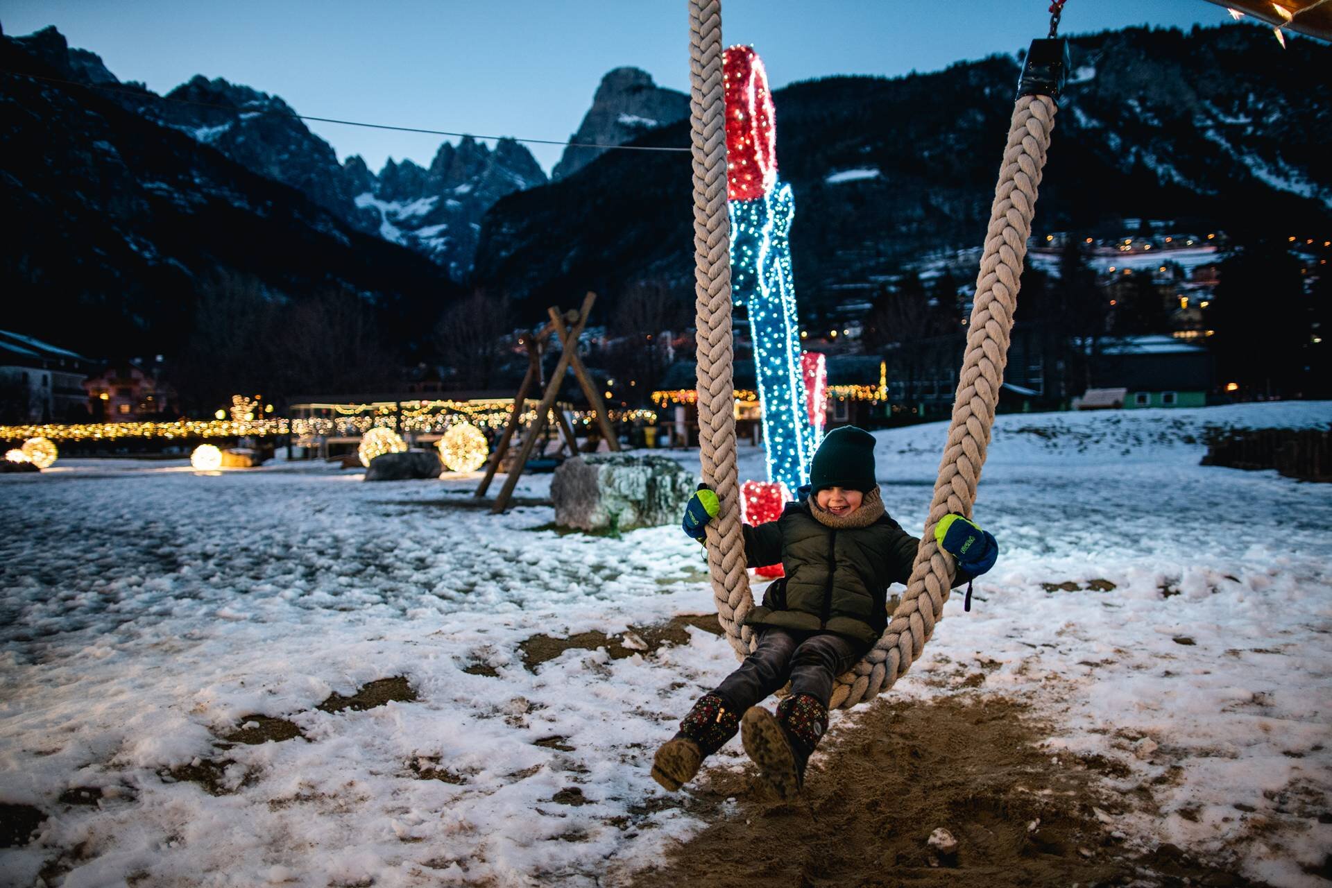 Un bambino vestito con una giacca verde e un berretto sta sorridendo mentre si dondola su una grande altalena di corda nel parco giochi all'aperto coperto di neve a Molveno sull'altopiano della Paganella. La scena è al crepuscolo, con luminarie natalizie sullo sfondo. Dietro si stagliano imponenti le Dolomiti di Brenta. | © Alice Russolo, 2022