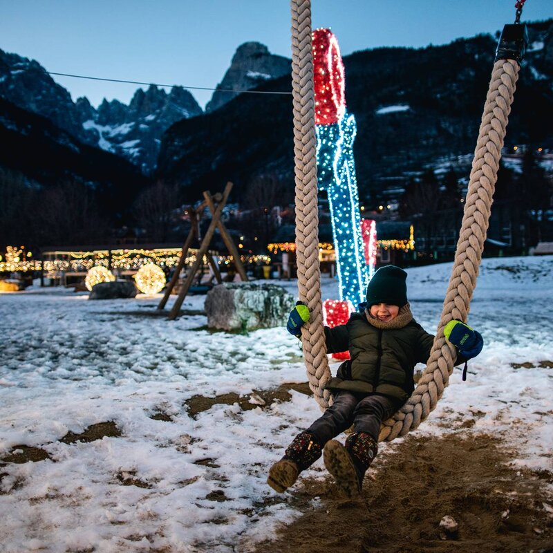 Un bambino vestito con una giacca verde e un berretto sta sorridendo mentre si dondola su una grande altalena di corda nel parco giochi all'aperto coperto di neve a Molveno sull'altopiano della Paganella. La scena è al crepuscolo, con luminarie natalizie sullo sfondo. Dietro si stagliano imponenti le Dolomiti di Brenta. | © Alice Russolo, 2022