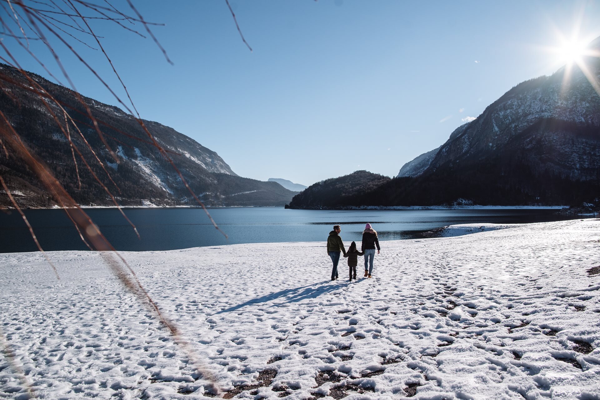 Drei Personen, von hinten gesehen, spazieren am schneebedeckten Ufer des Molvenosees entlang. Die Gruppe, bei der die mittlere Person von den beiden anderen an der Hand gehalten wird, geht an einem strahlenden Wintertag auf das ruhige Wasser zu, umgeben von imposanten Bergen. | © Alice Russolo, 2022