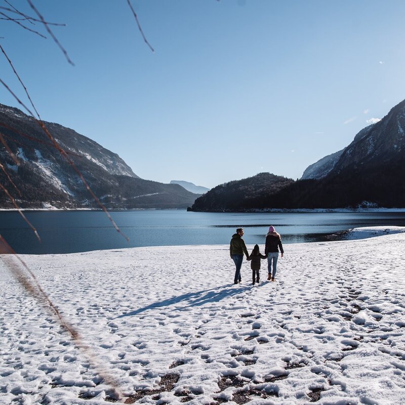 Tre persone, viste di schiena, passeggiano sulla riva innevata del Lago di Molveno. Il gruppo, con la persona al centro tenuta per mano dalle altre due, cammina verso l'acqua calma in una luminosa giornata invernale, circondato da imponenti montagne. | © Alice Russolo, 2022