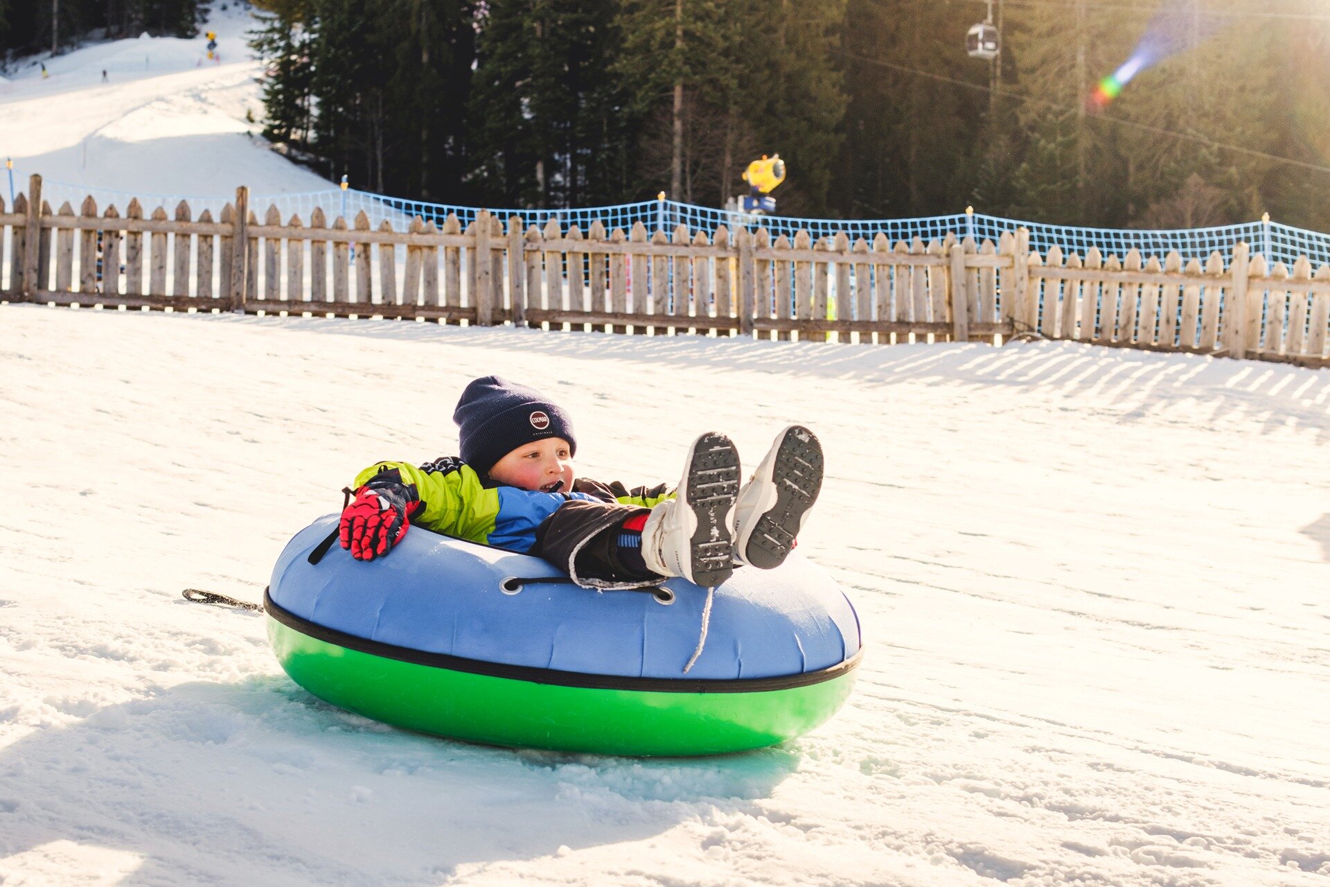A child slides down the snow on a colorful rubber tube (snow tubing) on a sunny winter day. In the background, framed by a wooden fence and safety nets, the snowy forest and a ski lift are visible. | © Filippo Frizzera, 2025