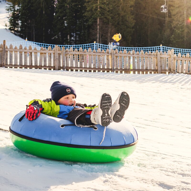 Un bambino scivola sulla neve a bordo di un gommone colorato (snow tubing) in una giornata di sole. Sullo sfondo, delimitato da una staccionata in legno e reti di protezione, si vede il bosco innevato e un impianto di risalita. | © Filippo Frizzera, 2025