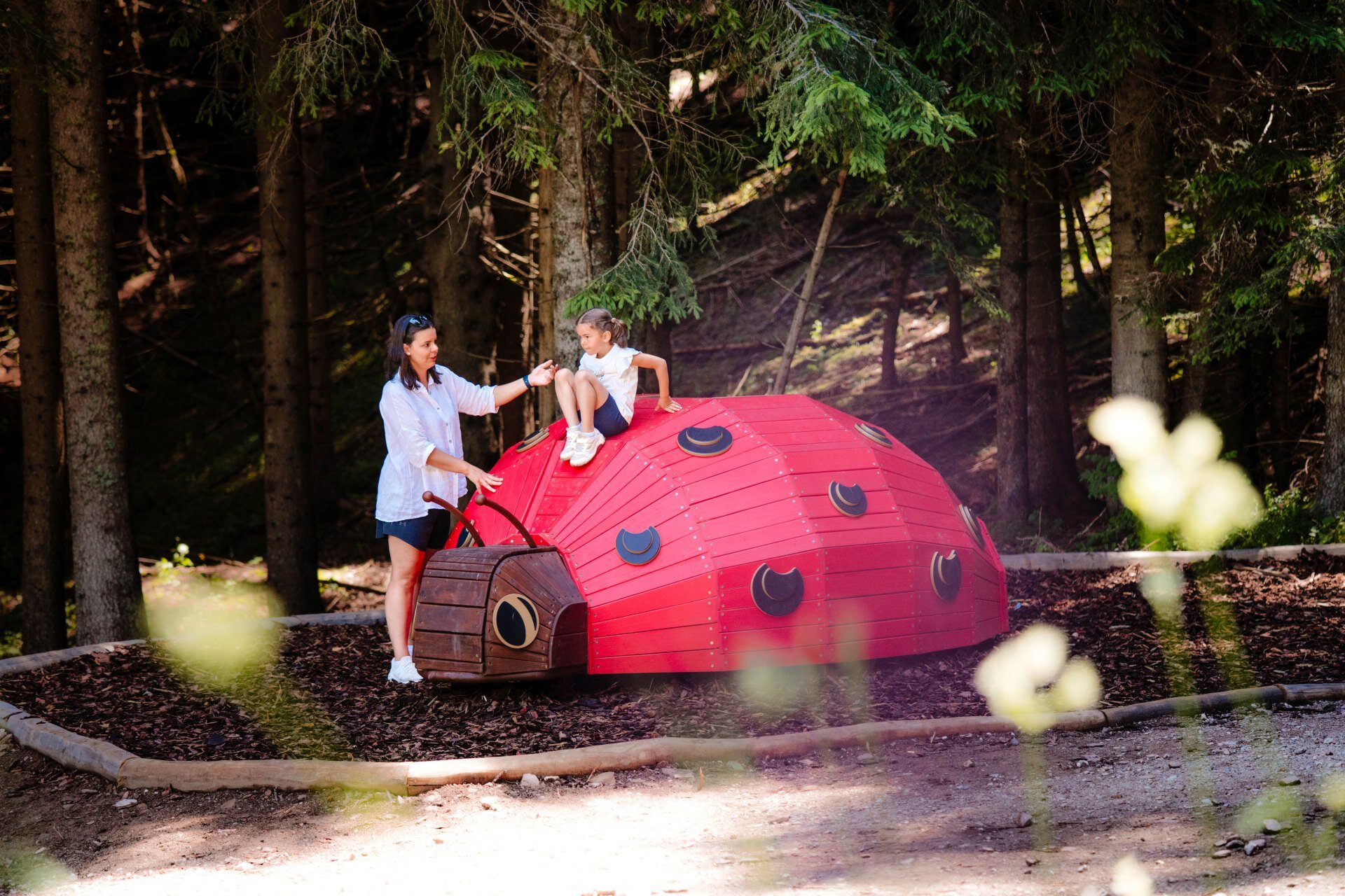 Una bambina è seduta sopra una grande scultura rossa a forma di coccinella, nel bosco della Paganella. Una donna in piedi accanto a lei le porge la mano per aiutarla. Entrambe sono circondate da alberi e vegetazione, su un terreno di corteccia. In primo piano, fiori sfocati. | © Mirko Perli, 2025