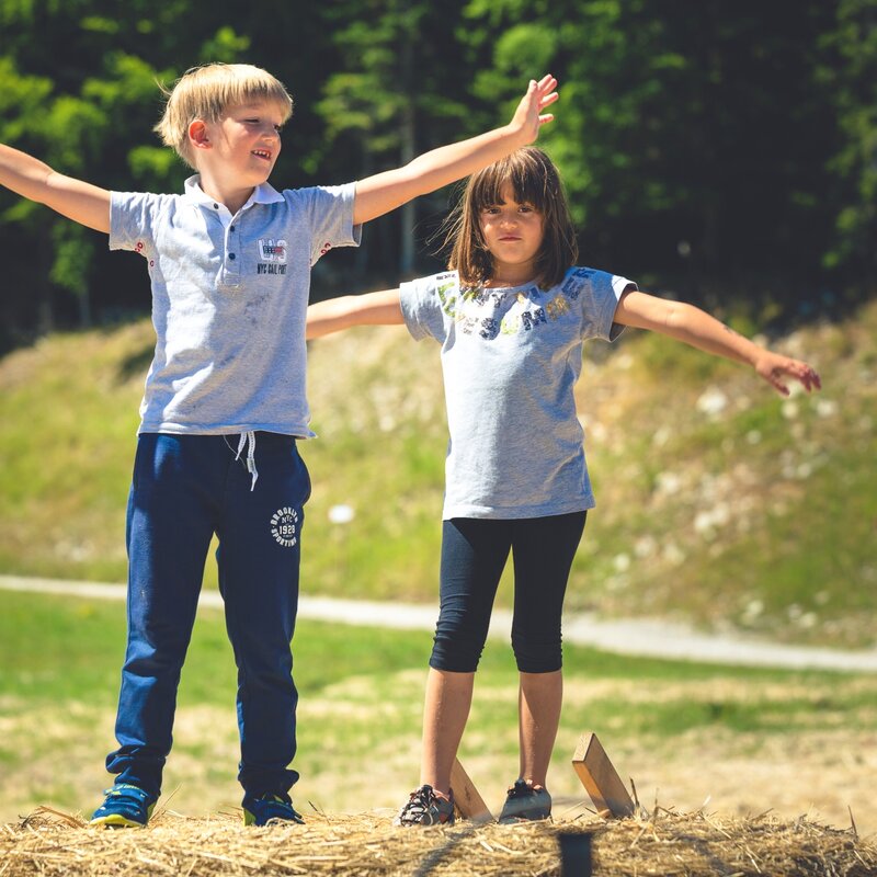 Due bambini, un maschio e una femmina, stanno in piedi su una balla di paglia rotonda con le braccia spalancate. Il ragazzo, biondo a sinistra, indossa una polo grigia e pantaloni blu. La ragazza, mora a destra, indossa una maglietta grigia e leggings scuri. Sono in un campo aperto con una collina boscosa e un sentiero sterrato sullo sfondo. | © Story Travelers, 2016