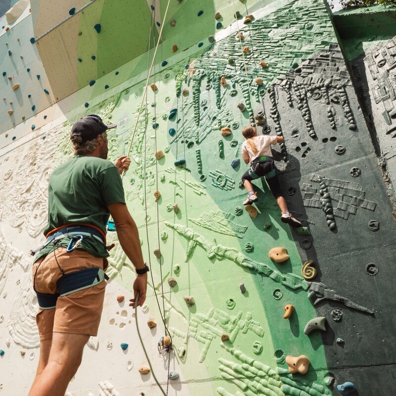 n uomo visto di spalle, con maglietta verde, pantaloncini beige e imbragatura, tiene in mano una corda di sicurezza ai piedi di una grande parete di arrampicata artificiale all'aperto. Più in alto sulla parete, una bambina con una t-shirt chiara e pantaloni scuri è intenta a salire, assicurata a una corda. La struttura presenta pannelli con diverse sfumature di verde, grigio e bianco, dotati di numerose prese colorate e texture a rilievo. | © Life Park, 2025