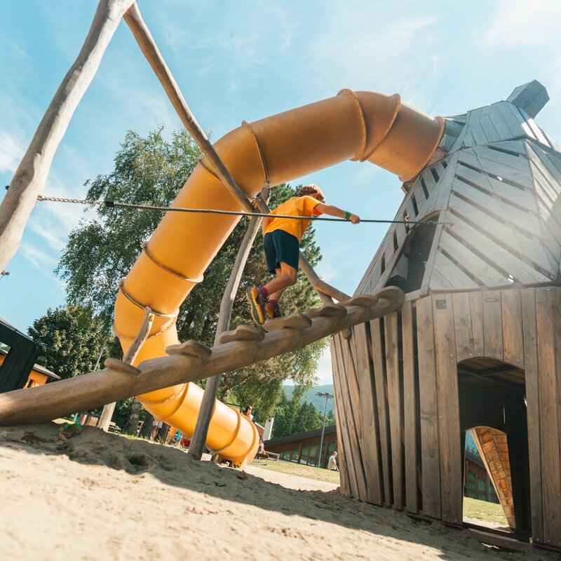 A child in an orange t-shirt climbs a slanted wooden log using a rope for balance, heading towards a large wooden play structure attached to an enclosed yellow tube slide. The ground is covered in light sand under a sunny blue sky. | © Andalo Gestioni, 2025