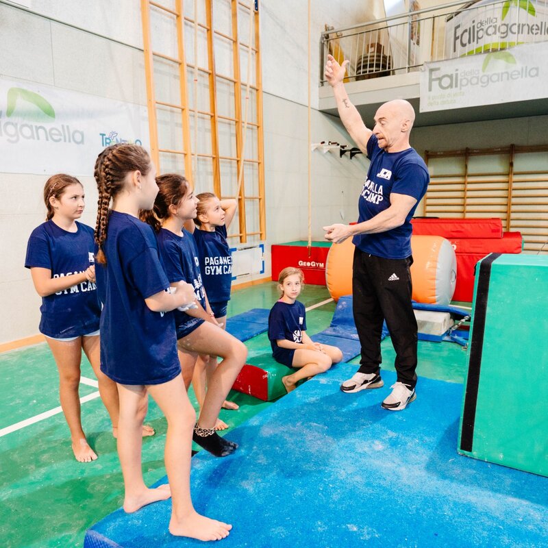 Jury Chechi, in piedi su un materasso blu, alza un braccio per dare indicazioni a un gruppo di cinque ginnaste ferme davanti a lui in ascolto. | © Mirko Perli, 2025