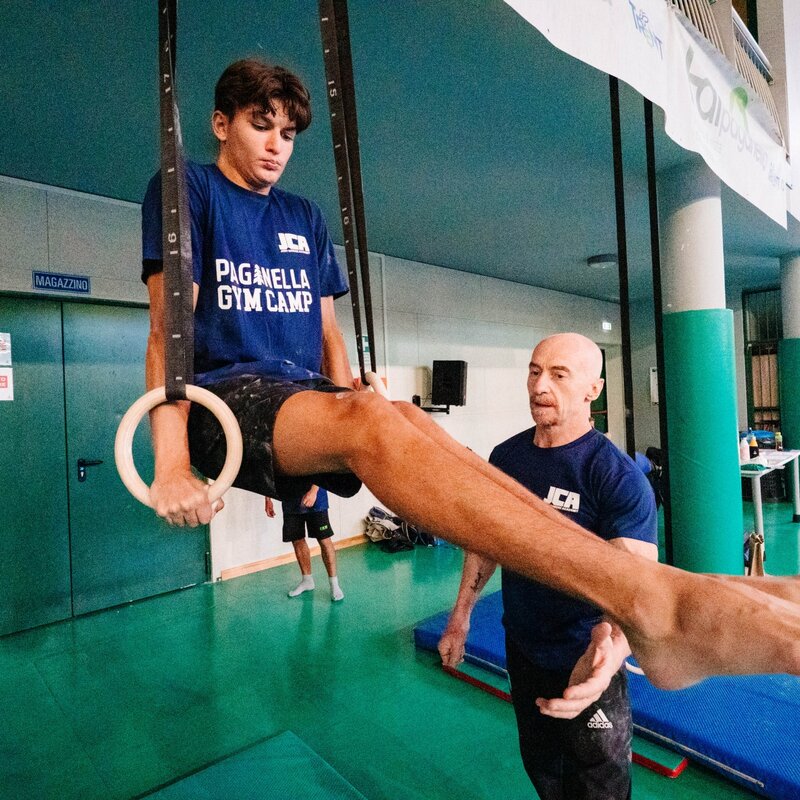 Un ginnasta esegue una tenuta a squadra (gambe tese parallele al suolo) agli anelli. Accanto a lui, Jury Chechi osserva la posizione tenendo le mani pronte per l'assistenza. | © Mirko Perli, 2025