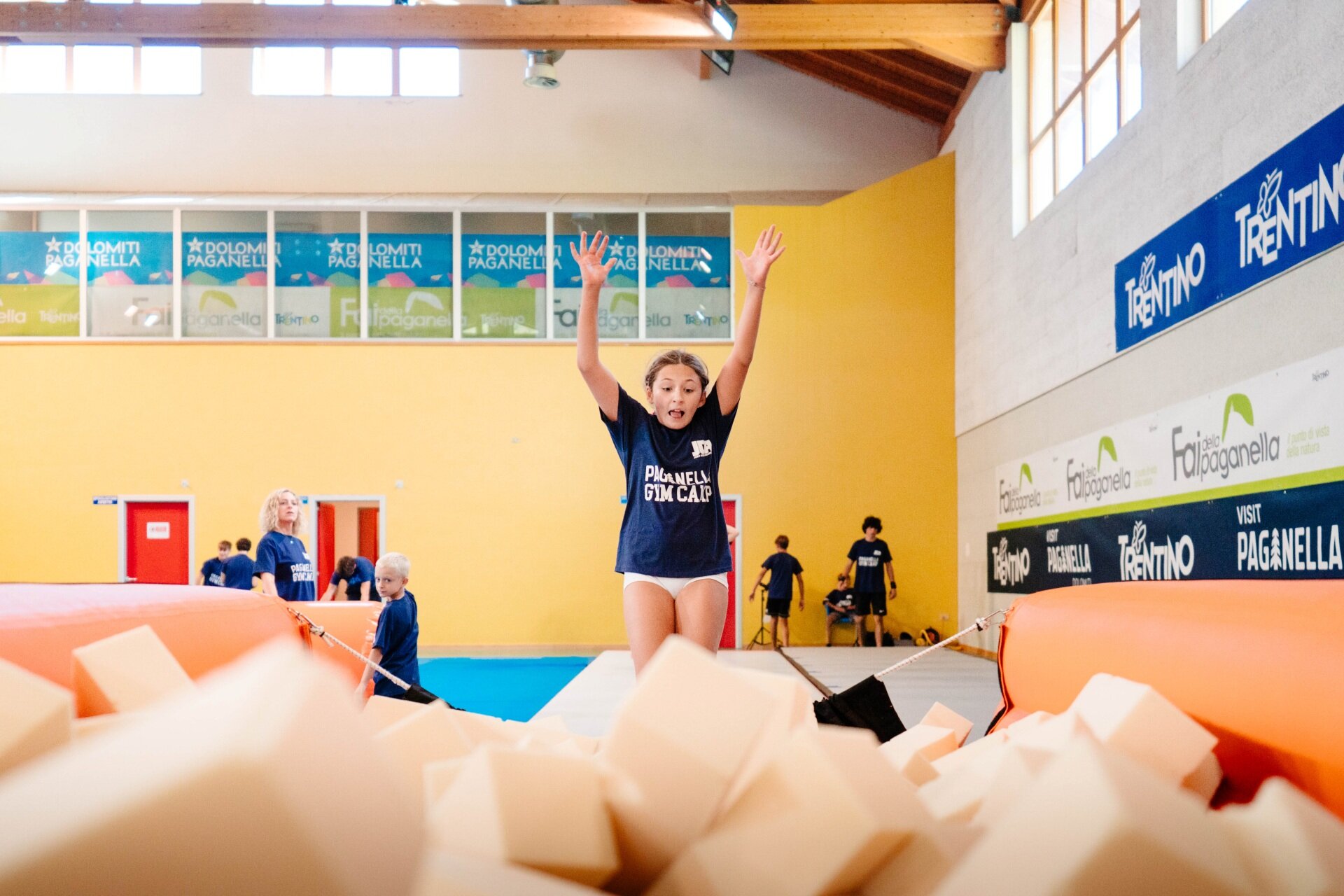 Una ginnasta è ripresa a mezz'aria, con le braccia alzate, mentre salta verso una grande vasca piena di cubi di gommapiuma. Alle sue spalle, sul pavimento azzurro della palestra, si trovano altri ginnasti in attesa e alcuni istruttori. | © Mirko Perli, 2025