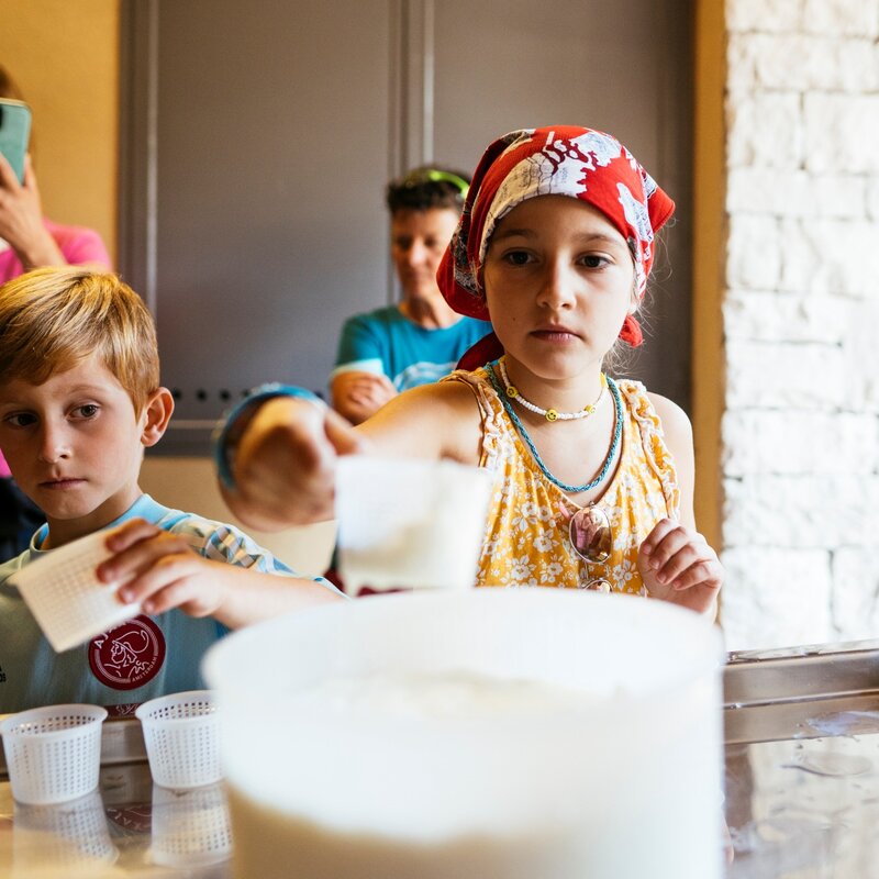 Due bambini partecipano a un laboratorio di caseificazione presso Malga Fai. In primo piano, una bambina con bandana rossa e un bambino con maglia azzurra riempiono piccoli stampi forati bianchi attingendo da un secchio di cagliata fresca. Sullo sfondo, alcune persone osservano l'attività in un ambiente rustico con pareti in pietra. | © Giovanni Danieli, 2021