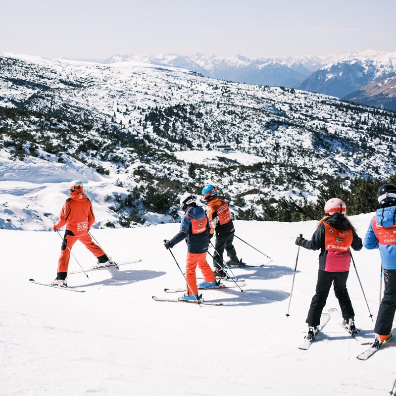 A group of young skiers wearing red bibs follows a ski instructor dressed in red down a gentle slope. The group is viewed from behind as they ski towards the valley, with a wide panorama of snow-capped mountains and scattered vegetation in the background. | © Filippo Frizzera, 2025