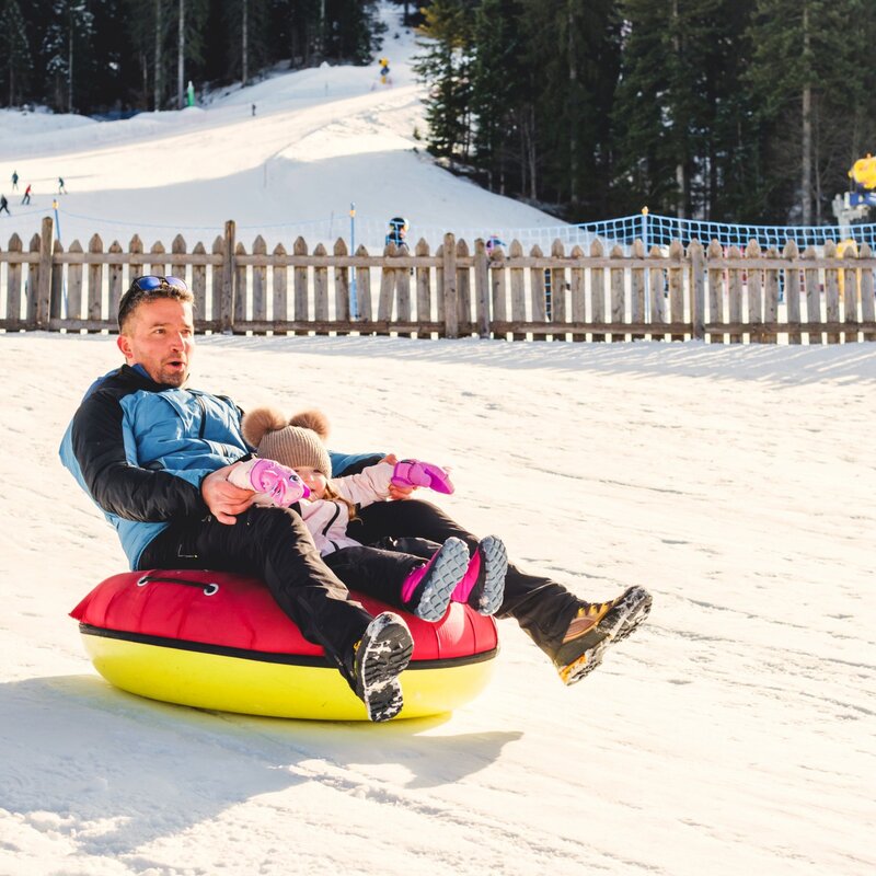 Ein Mann und ein kleines Mädchen rutschen gemeinsam auf einem rot-gelben Reifen einen sonnigen, verschneiten Hang hinunter. Hinter ihnen befinden sich ein Holzzaun und Sicherheitsnetze, während im Hintergrund eine Skipiste mit anderen Skifahrern und ein Nadelwald zu sehen sind. | © Filippo Frizzera, 2025