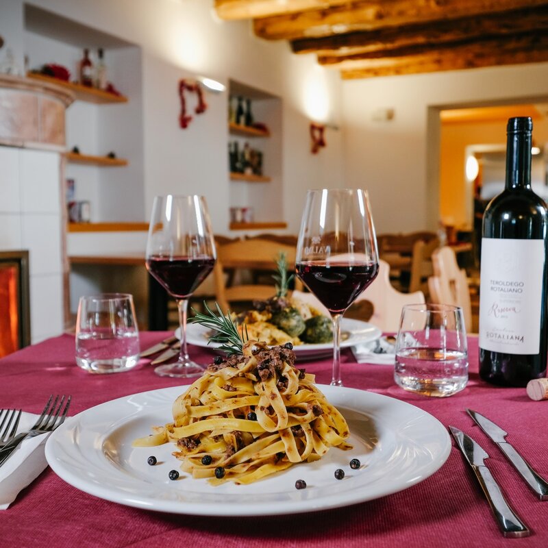 A table set in front of the fireplace at Malga Terlaga. In the foreground a plate of tagliatelle with mushrooms and two glasses of Teroldego Rotaliano, a red wine. A second dish can be glimpsed in the background. | © Multiweb, 2019
