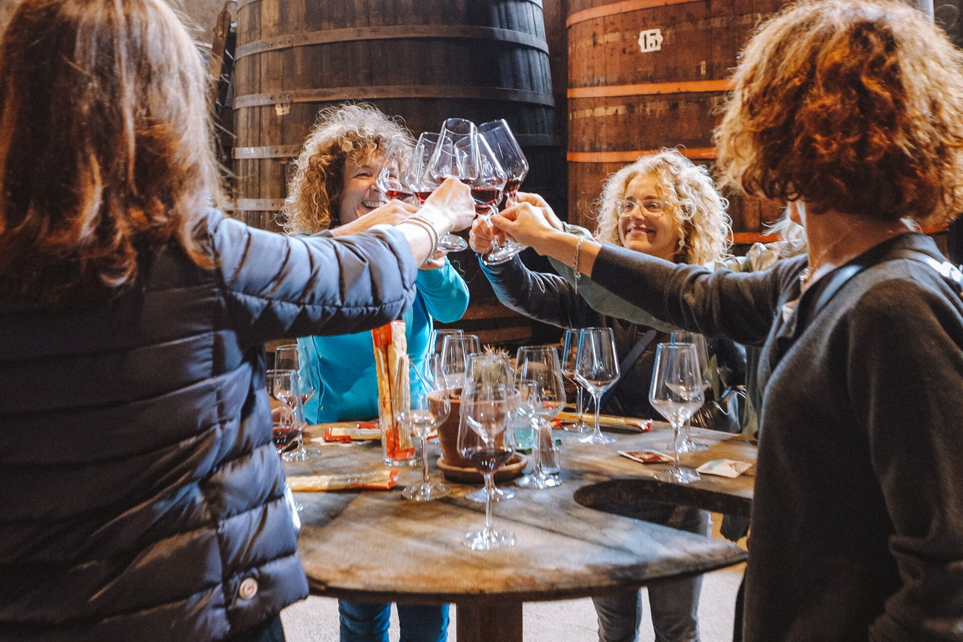A group of smiling women makes a toast with glasses of red wine, gathered around a wooden table in a wine cellar. The scene, taking place at Cantina Martinelli in Piana Rotaliana, captures a convivial moment during a wine tasting, with large oak barrels visible in the background. | © Nicola Cagol, 2021