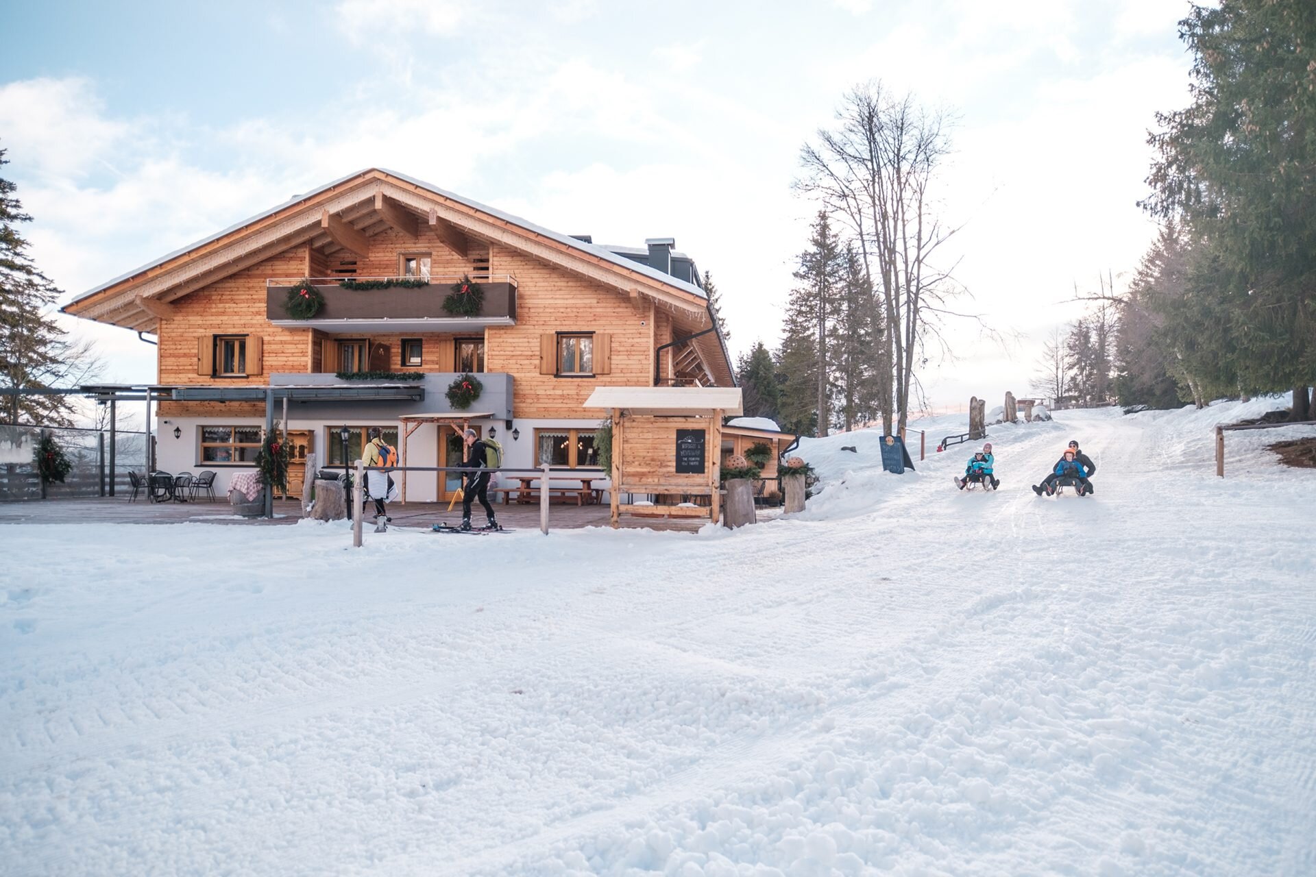 Winterszene. Kinder und Erwachsene rodeln im Vordergrund einen verschneiten Hang hinunter, mit der charakteristischen Holzhütte Rifugio La Montanara im alpinen Stil im Hintergrund. | © Filippo Frizzera, 2022