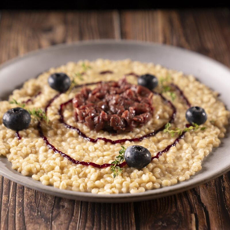 Piatto di orzotto cremoso con tartare di cervo al centro, decorato con una spirale di riduzione al Teroldego Rotaliano, mirtilli freschi e timo. | © Stefano Caffarri, 2025