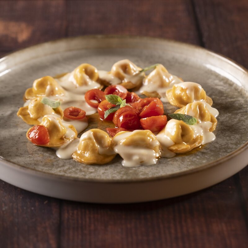 Tortelli ripieni di trota igp e disposti a cerchio conditi con crema di formaggio, pomodorini rossi al centro e foglioline di erbe aromatiche. | © Stefano Caffarri, 2025