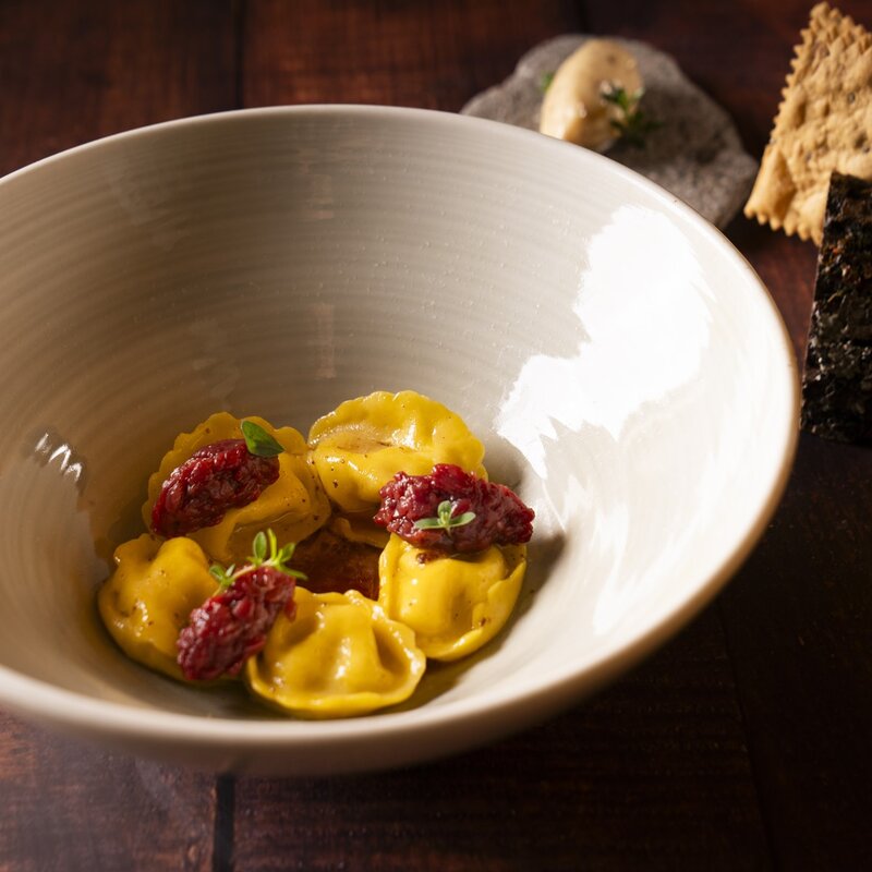 Ravioli ripieni al sedano rapa con tartare di cervo, serviti in infuso di sambuco con cracker di segale e burro al fieno sullo sfondo. | © Stefano Caffarri, 2025