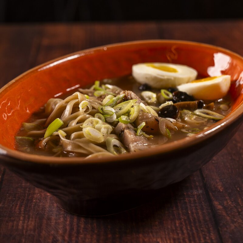 Tegame arancione con zuppa di ramen, uovo sodo a metà, spezzatino di maiale e cipollotto fresco. | © Stefano Caffarri, 2025