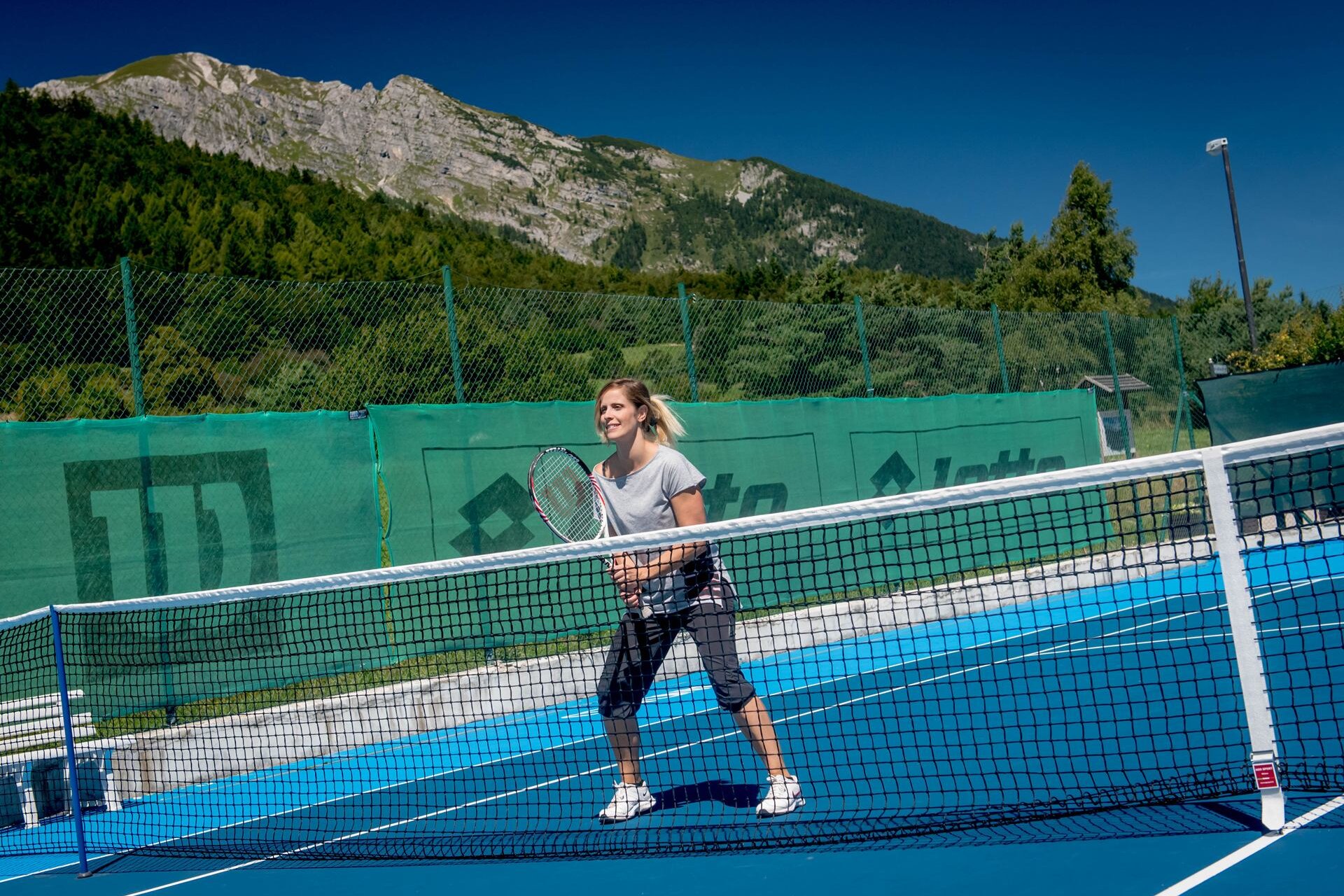 Eine blonde Frau mit gebeugten Knien und einem Tennisschläger in der Hand wartet auf dem Tennisplatz von Andalo Life auf ihren Aufschlag. Im Hintergrund sind die Brenta-Dolomiten zu sehen | © Matteo De Stefano, 2016