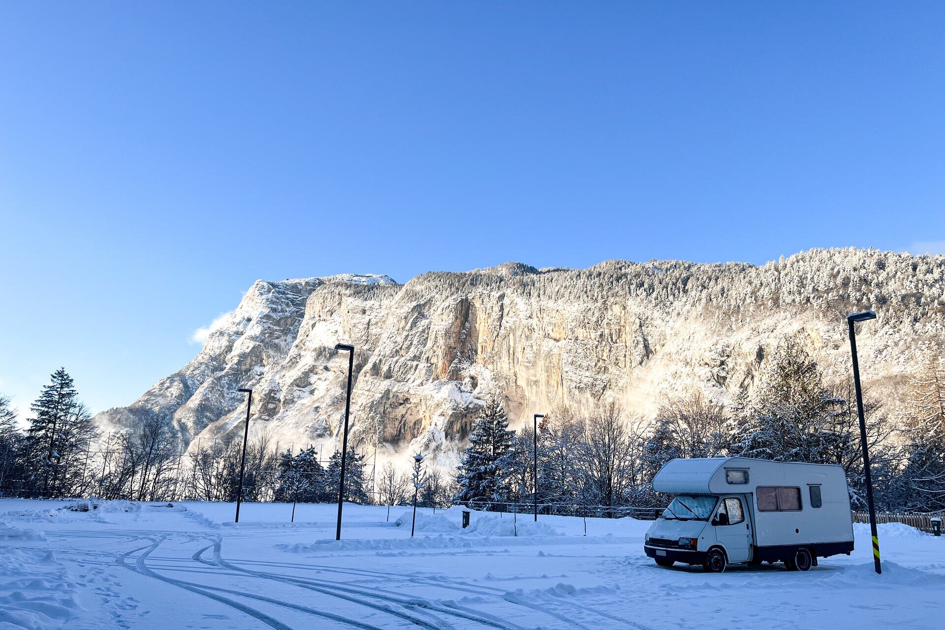 An einem sonnigen Wintertag steht ein weißer Wohnwagen auf einem vollständig verschneiten Rastplatz. Im Vordergrund sind frische Reifenspuren im Schnee zu sehen. Im Hintergrund dominiert der große Berg Paganella die Szene, dessen Felswand teilweise mit Schnee und Bäumen bedeckt ist. Der Himmel ist klar und blau. | © Fai Vacanze, 2023