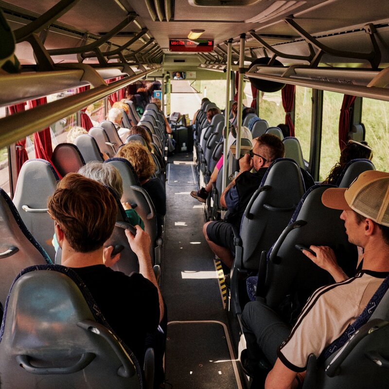 Vista dall'interno di un autobus pieno di passeggeri seduti, inquadrati dal fondo verso la parte anteriore. In primo piano a destra, un ragazzo indossa un cappellino con visiera. | © Origami, 2026