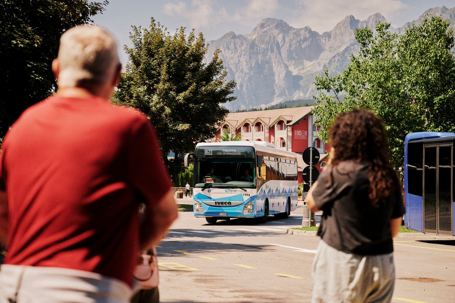 Un autobus di linea bianco e azzurro con l'insegna "TRENTO" viaggia verso l'osservatore su una strada asfaltata. In primo piano, due persone sfocate viste di spalle. Sullo sfondo, alberi, un edificio e alte cime montuose. | © Origami, 2026