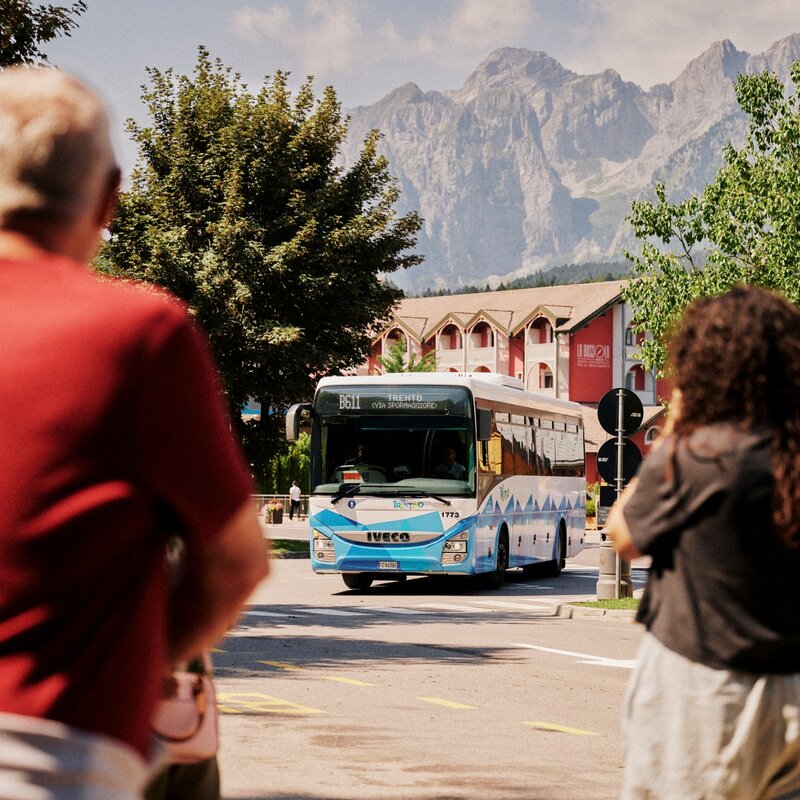 Un autobus di linea bianco e azzurro con l'insegna "TRENTO" viaggia verso l'osservatore su una strada asfaltata. In primo piano, due persone sfocate viste di spalle. Sullo sfondo, alberi, un edificio e alte cime montuose. | © Origami, 2026