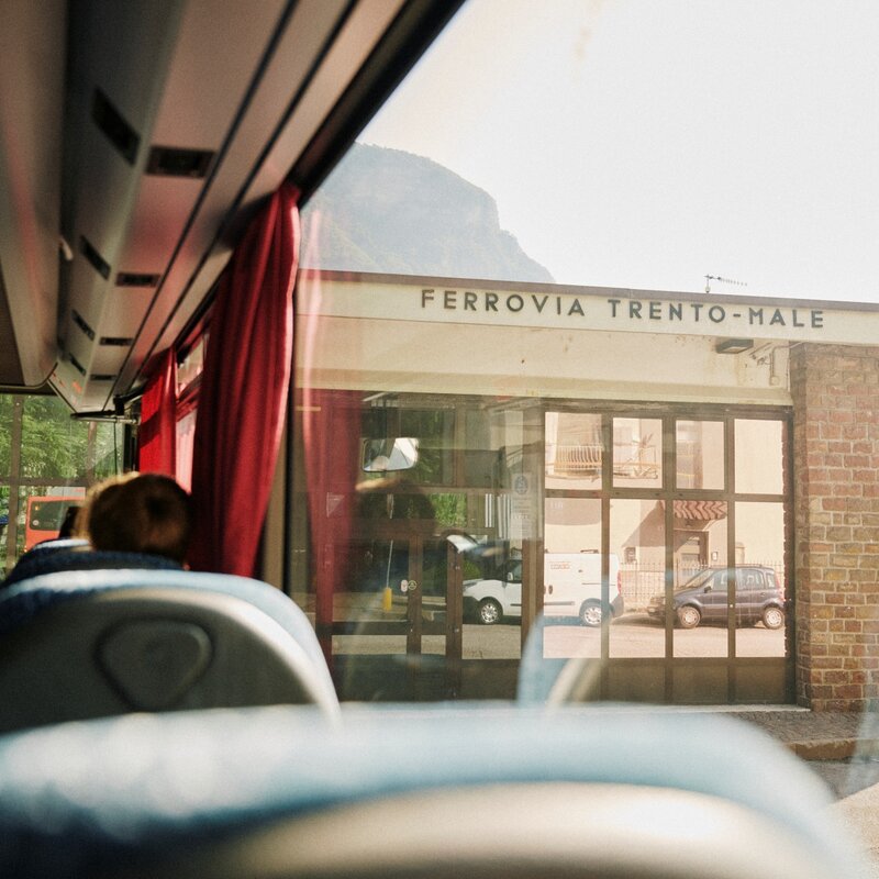 : Vista dall'interno di un autobus verso un edificio in mattoni con l'insegna "FERROVIA TRENTO-MALE". In primo piano, le teste dei passeggeri seduti. | © Origami, 2026