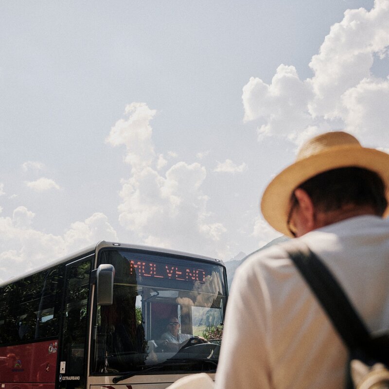 Un autobus rosso con l'insegna luminosa "MOLVENO" si avvicina sulla strada. In primo piano, sfocata, una persona vista di spalle con un cappello di paglia. Sullo sfondo, montagne e cielo nuvoloso. | © Origami, 2026