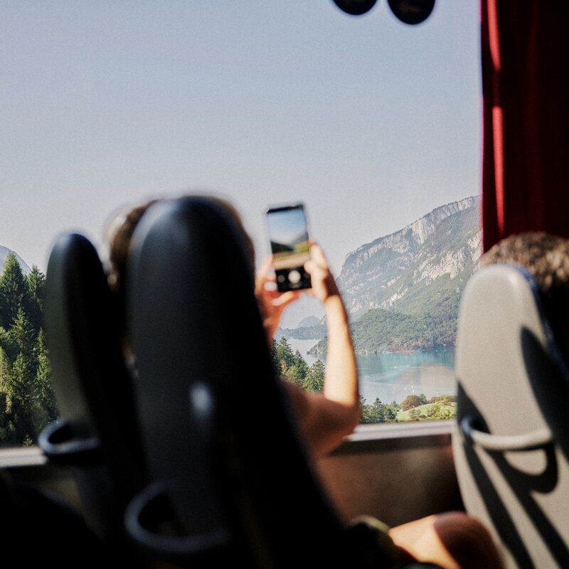 Vista dall'interno di un autobus dove un passeggero fotografa il paesaggio dal finestrino con uno smartphone. All'esterno si vede un lago circondato da montagne boscose e pareti rocciose. | © Origami, 2026